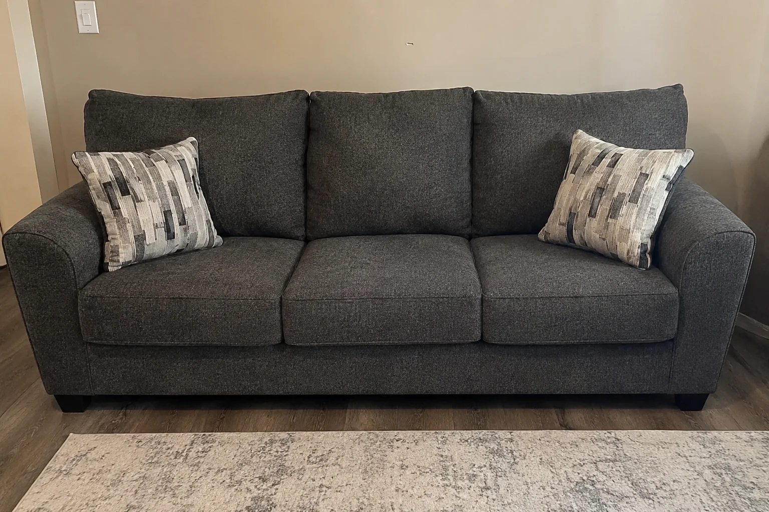 Three-seater sofa with dark gray upholstery, rolled armrests, black feet, and two patterned throw pillows.